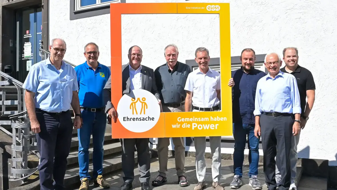 Acht Männer stehen vor dem Eingang eines Rathauses und halten einen orangenen Rahmen mit der Aufschrift "evm Ehrensache". Sie lächeln freundlich.