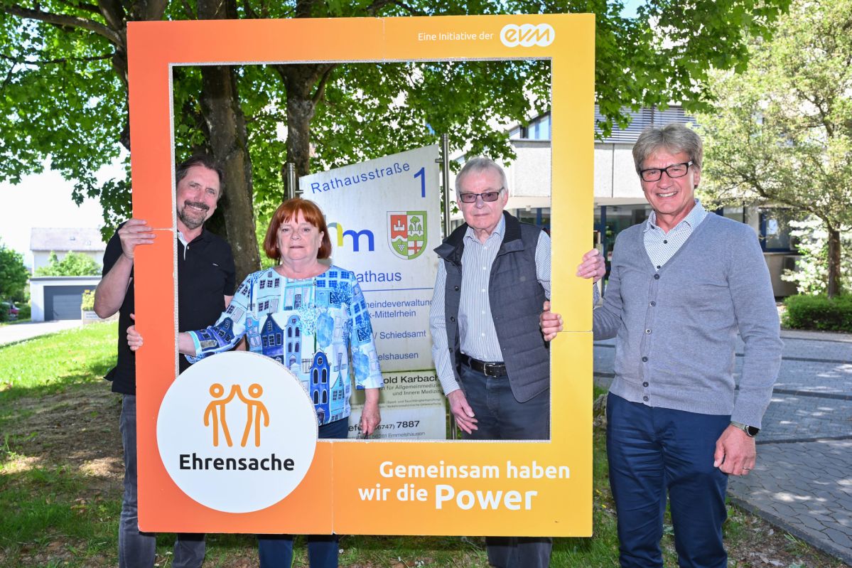 Drei Männer und eine Frau lächeln freundlich und halten einen orangenen Rahmen mit der Aufschrift "Ehrensache".