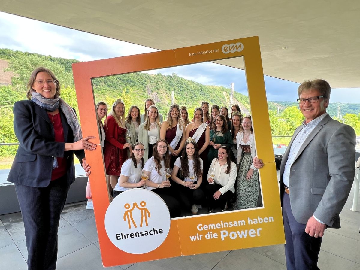Ehrensache Rhein-Mosel Eine Gruppe von jungen Frauen mit Weingläsern steht hinter einem orangenen Rahmen mit der Aufschrift "Ehrensache". Der Rahmen wird gehalten von einer Frau und einem Mann.