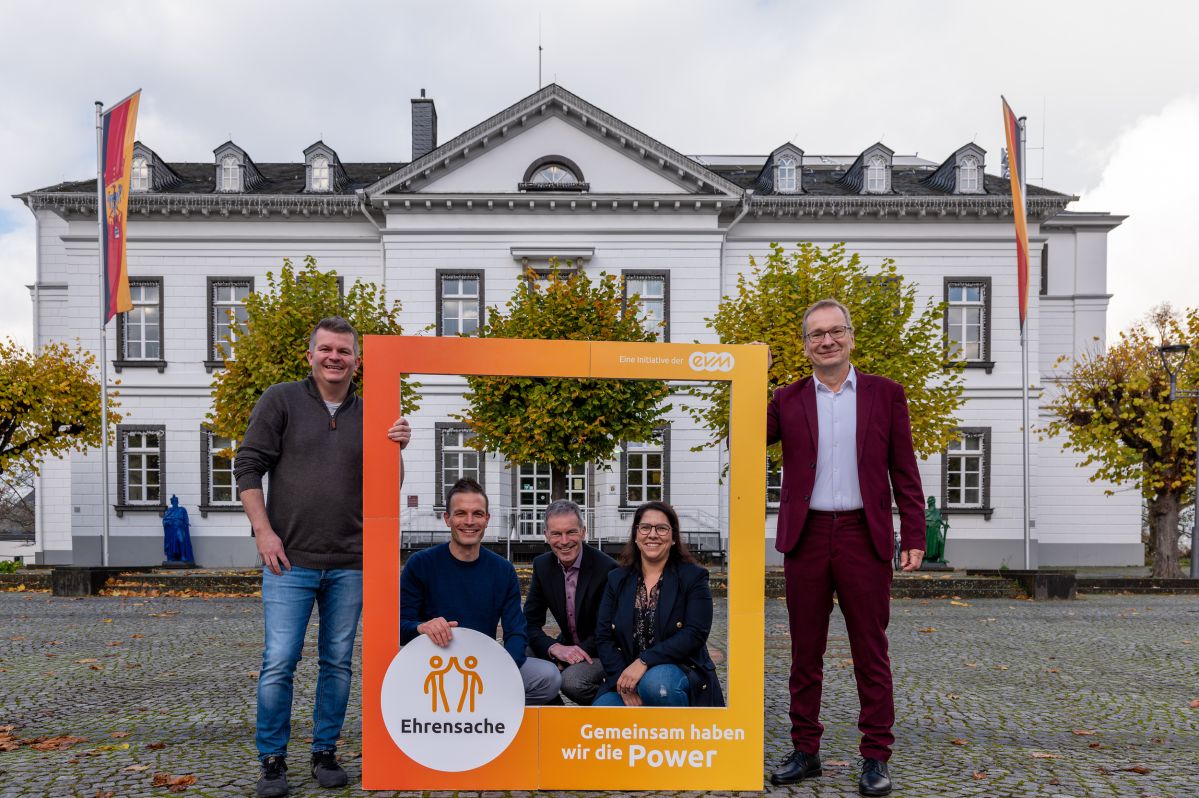 Vier Männer und eine Frau stehen vor einem Amtsgebäude und halten einen orangenen Rahmen mit der Aufschrift "evm Ehrensache". Sie lächeln freundlich.