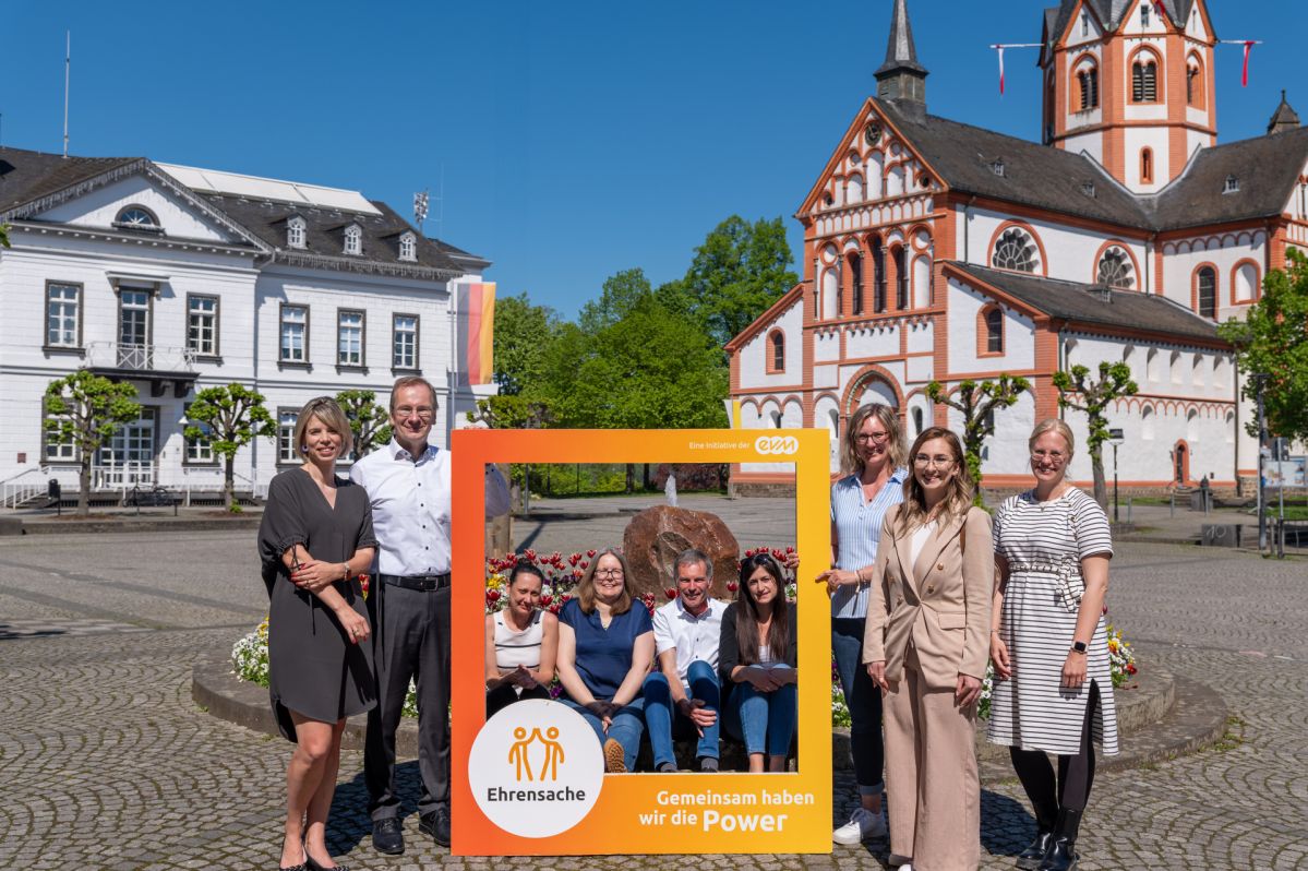 Sieben Frauen und zwei Männer stehen bzw. sitzen auf einem Platz und halten einen orangenen Rahmen mit der Aufschrift "evm Ehrensache". Sie lächeln freundlich. Im Hintergrund ist ein Gebäude und eine Kirche zu sehen.
