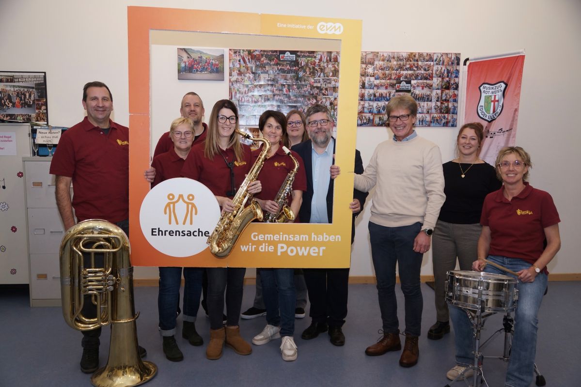 Eine Gruppe an Musikern mit Instrumenten stehen in einem Raum und halten einen orangenen Rahmen mit der Aufschrift "evm Ehrensache". Sie lächeln freundlich.