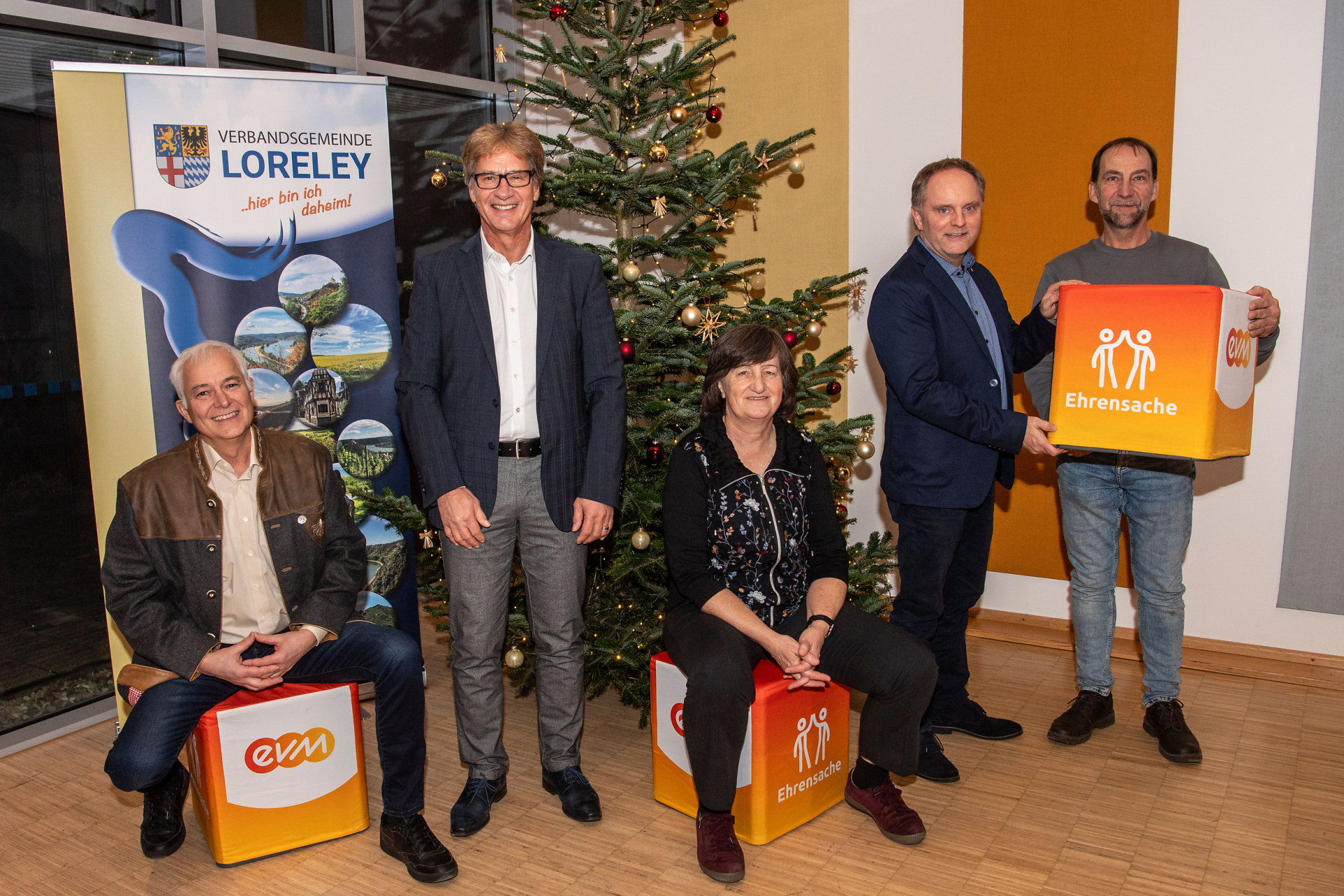 Eine Personengruppe vor einem Weihnachtsbaum präsentiert „evm“‑ und „Ehrensache“-Würfel, neben einem Banner der Verbandsgemeinde Loreley.
