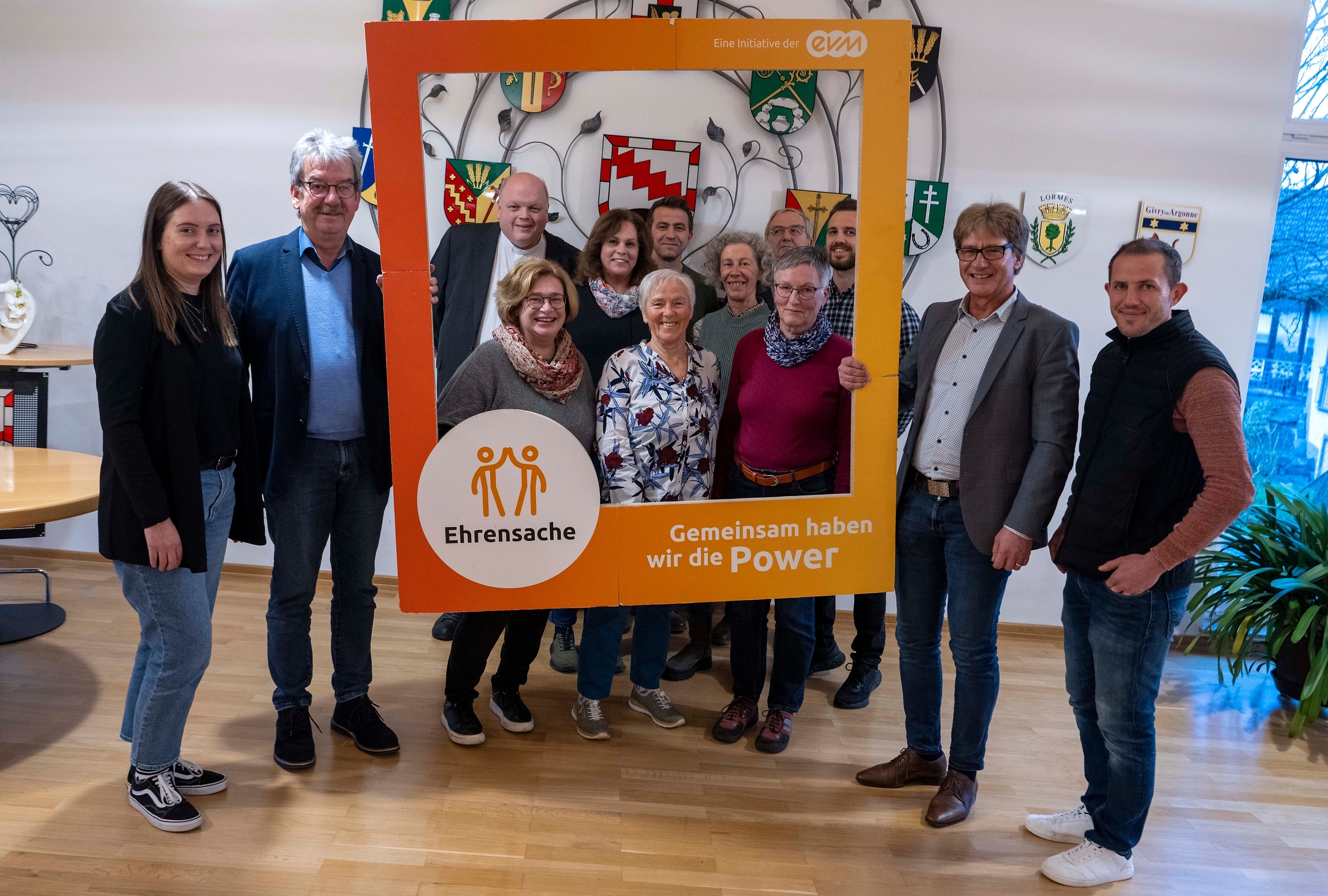 Eine Gruppe von 12 Personen steht in einem Raum und posiert mit einem großen, orangefarbenen Rahmen. Auf dem Rahmen steht: „Ehrensache - Gemeinsam haben wir die Power“. Im Hintergrund sind Wappen sichtbar.