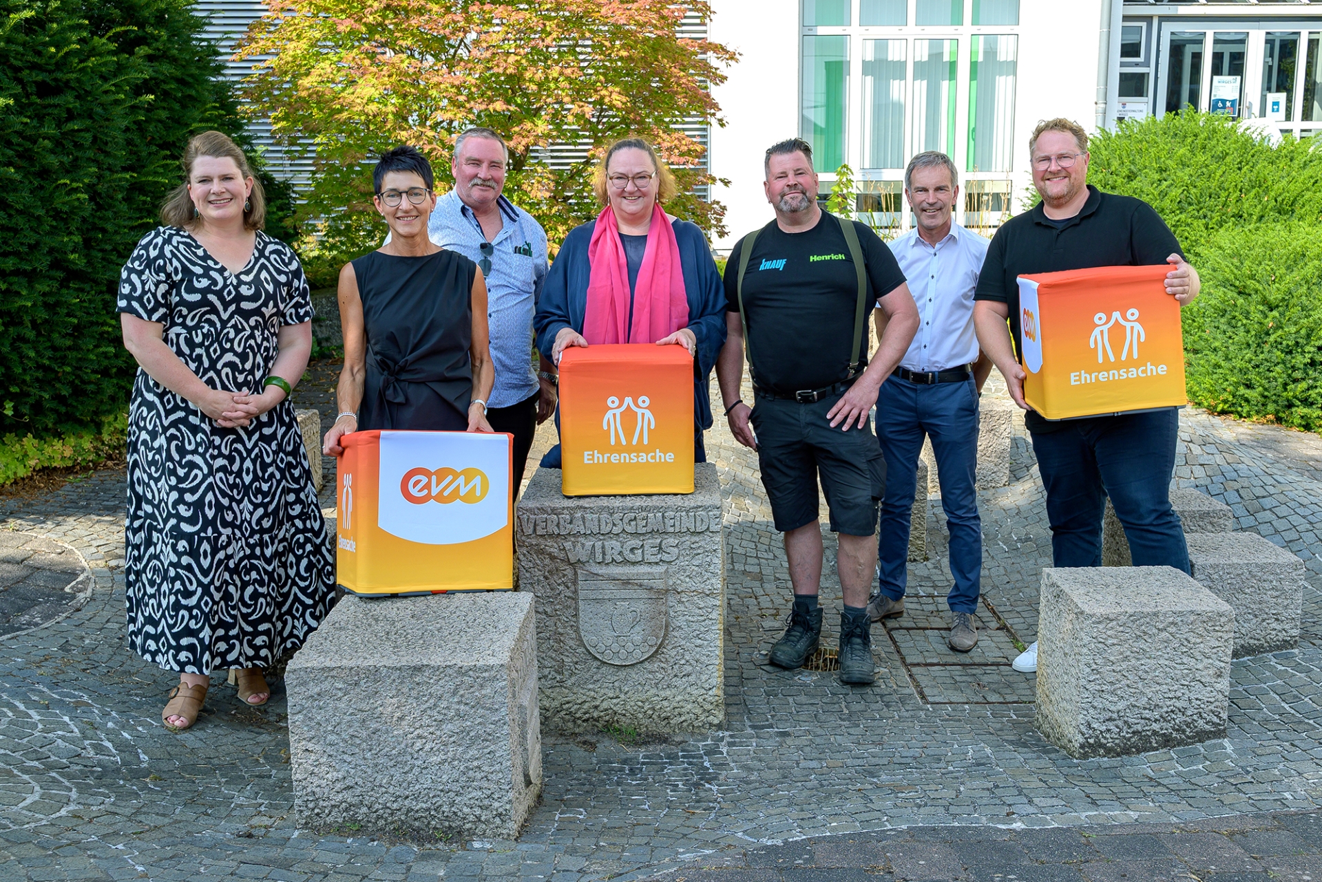 Sieben Erwachsene stehen freundlich lächelnd zusammen. Vor ihnen stehen orangene evm Ehrensache Würfel.
