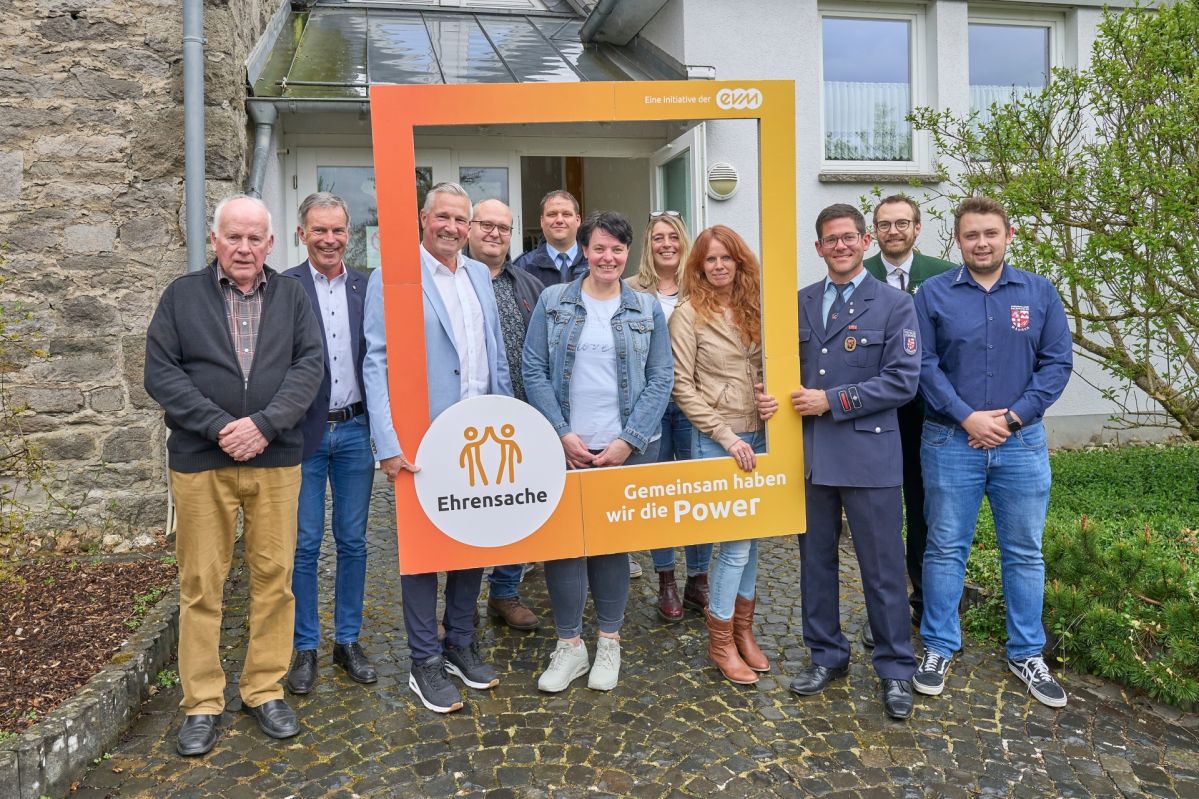 evm-Ehrensache - Verbandsgemeinde Wallmerod 2025 11 Personen stehen freundlich lächelnd vor einem Gebäude. Sie halten einen orangenen Rahmen mit der Aufschrift "Ehrensache".