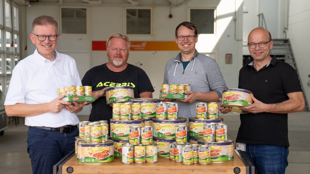 Vier Männer stehen vor einem Tisch auf dem Konserven gestapelt sind und lächeln freundlich.