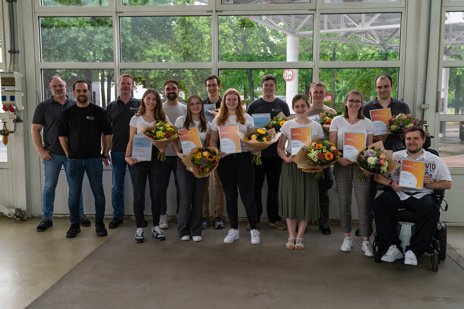 Eine Gruppe junger Menschen steht in einer Halle und lächelt freundlich. Sie halten Zertifikate in der Hand und Blumensträuße.