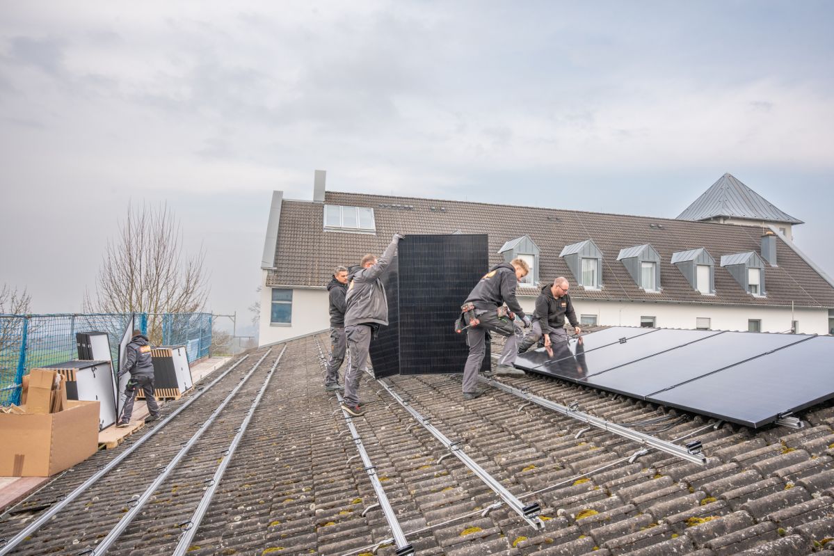 evmS Kloster-30 Vier Handwerksmonteure tragen ein Solarpanel auf einem Dach. Im Hintergrund sind weiter Panel erkennbar, sowie ein Haus. Der Himmel ist bewölkt.