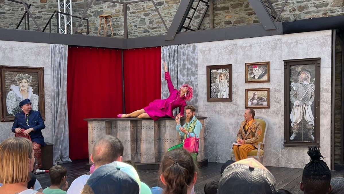 Eine Theaterbühne mit einem roten Vorhang und grauen Wänden. An den Wänden hängen mehrere Gemälde. Vier bunt gekleidete Schauspieler sind auf der Bühne. Im Vordergrund sieht man die Hinterköpfe von Personen im Publikum.