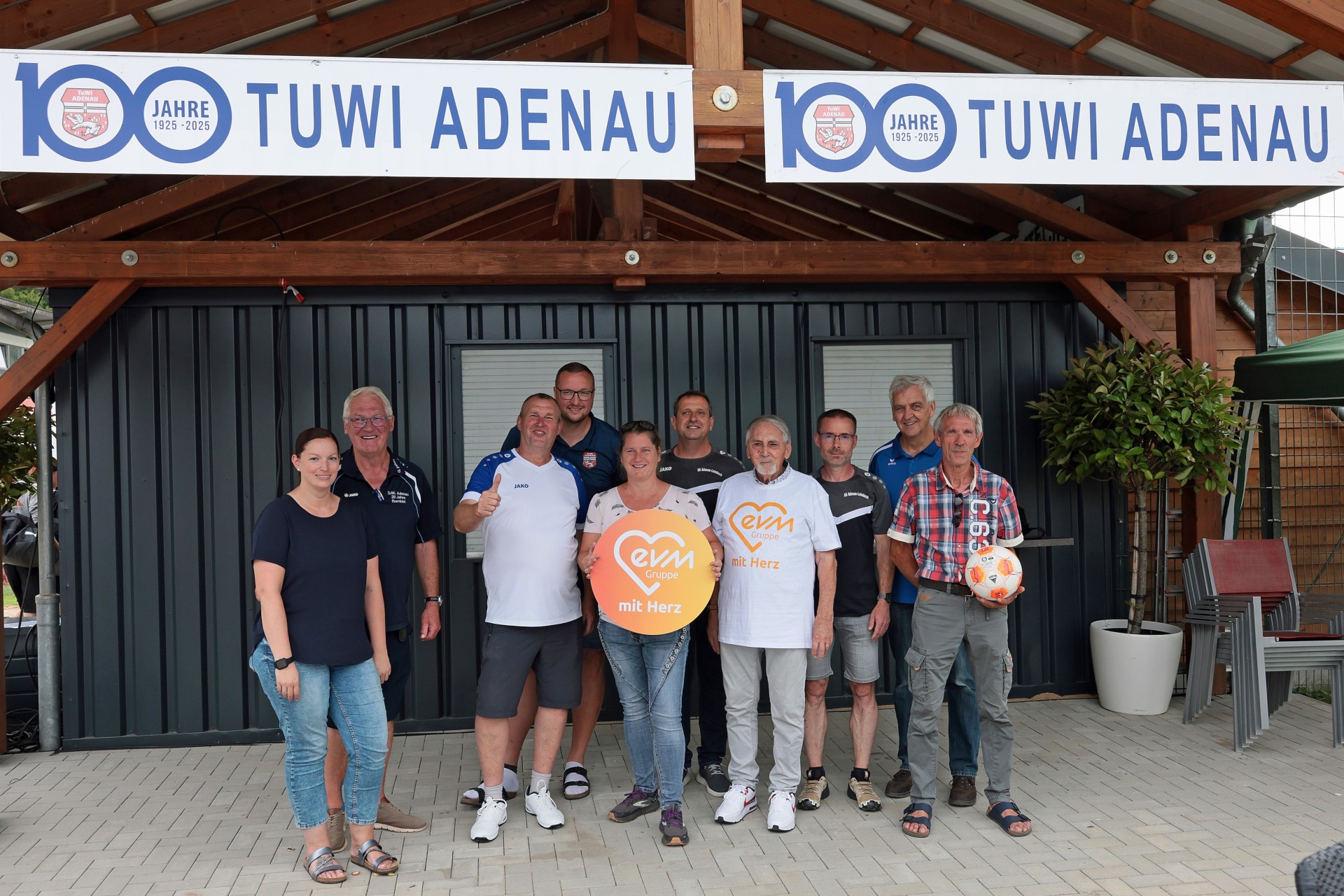Zwei Frauen und Acht Männer stehen vor einem Verkaufsstand und lächeln freundlich. Sie halten ein orangenes Schild mit Aufschrift "evm mit Herz".