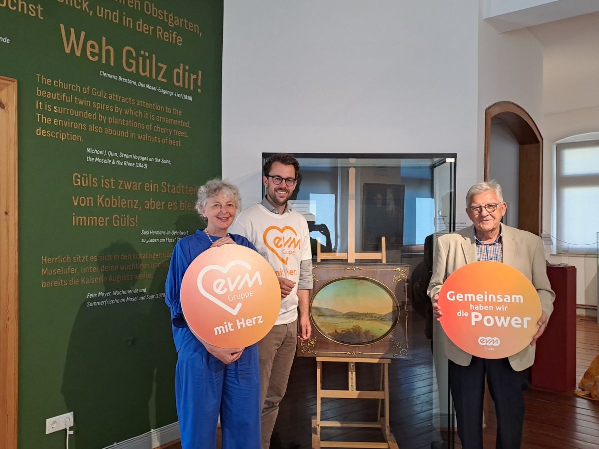 Eine Frau und zwei Männer stehen in einem Museum vor einem Gemälde. Sie halten kreisförmige, orangene Schilder mit den Aufschriften "evm mit Herz" und "Gemeinsam haben wir die Power".