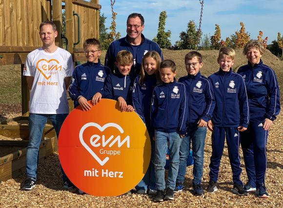 Mehrere Kinder in dunkelblauen Jacken und zwei Männer stehen auf einem Spielplatz und lächeln freundlich. Sie halten ein orangenes kreisförmiges Schild mit der Aufschrift "evm mit Herz".