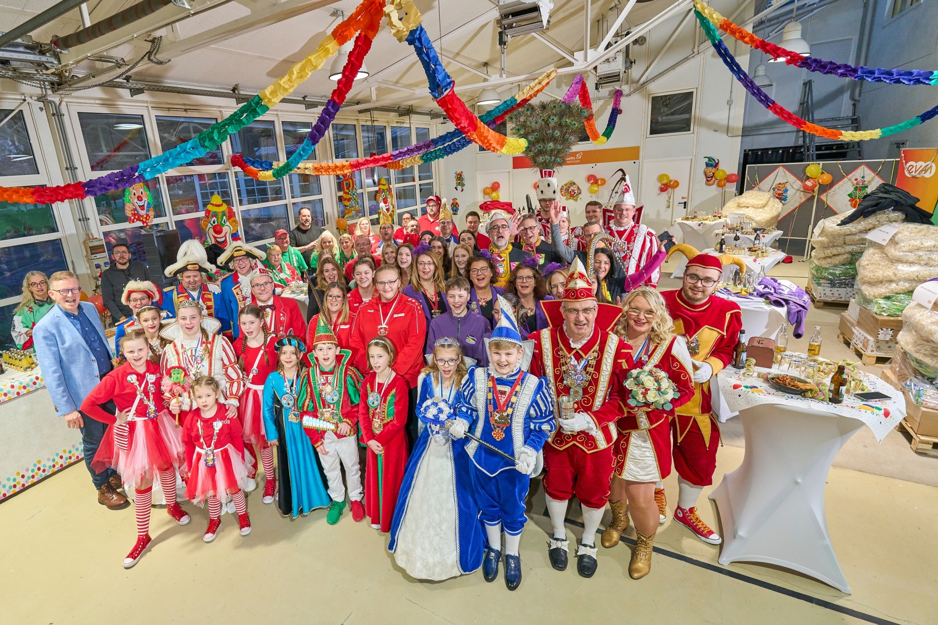 evm-Gruppe Palettenparty 2025 Eine große Gruppe Menschen in bunten Karnevalskostümen lächelt und posiert in einem festlich dekorierten Raum mit Luftschlangen und Snacks auf einem Tisch im Hintergrund.
