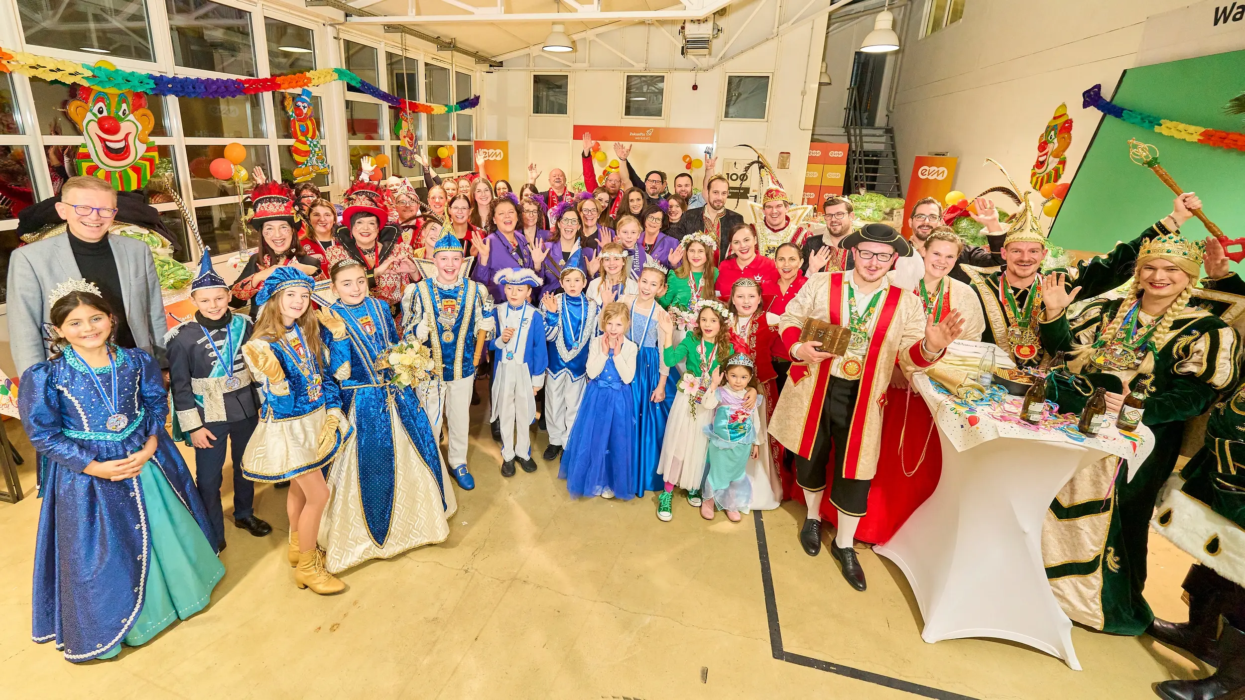 Eine große Gruppe von Menschen in farbenfrohen Kostümen posiert lächelnd in einem festlich geschmückten Raum mit Girlanden, Luftballons und Clown-Dekorationen, vermutlich bei einer Karnevalsfeier.