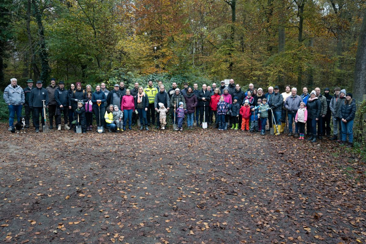 Baumpflanzaktion KO-01 Eine große Gruppe an Erwachsenen und Kindern stehen mit Schaufeln in einem Wald.