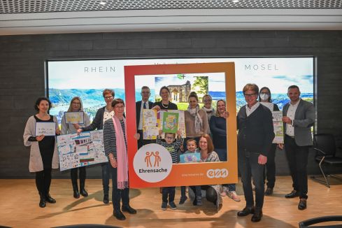 Eine Gruppe von Menschen, darunter zwei Kinder, hält bunte Zeichnungen und einen großen, rot-orangefarbenen Bilderrahmen mit der Aufschrift „Ehrensache, Eine Initiative der evm“. Im Hintergrund Landschaftsbilder.
