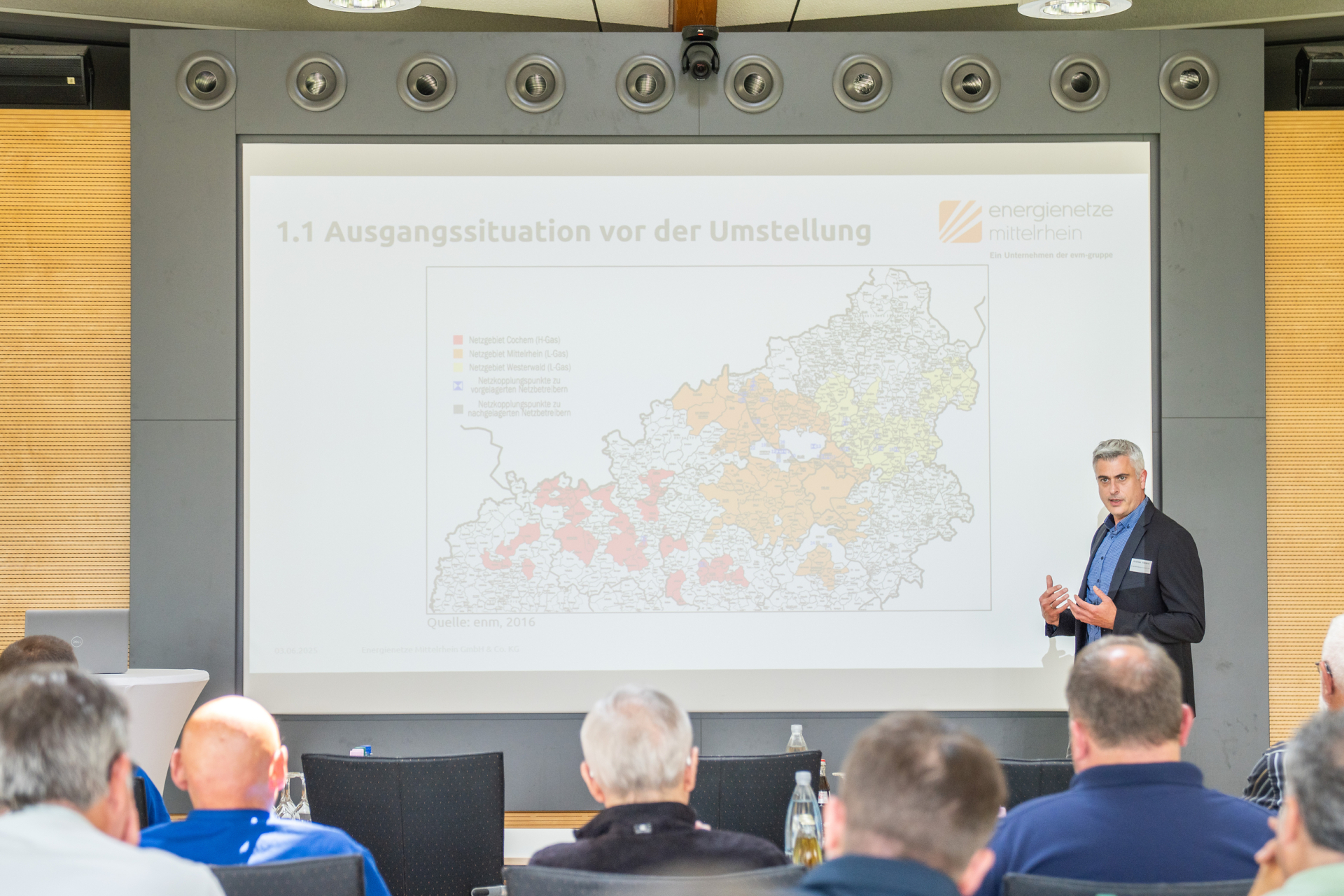 Ein Mann steht vor einem großen Bildschirm auf dem eine Karte zu sehen ist. Er steht vor einem Publikum und ist professionell gekleidet.