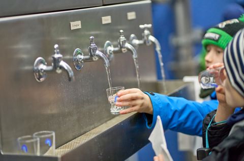 Zwei Jungen stehen vor einem großen Wasserspender mit vier Hähnen und halten Wassergläser da drunter.