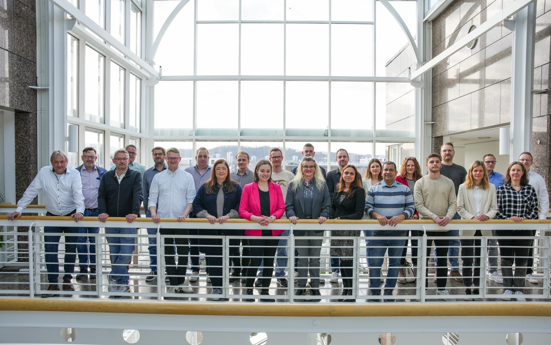 24 Menschen stehen freundlich lächelnd auf einer Balustrade. Im Hintergrund ist ein großes Fenster mit einer verschwommenen urbanen Landschaft zu erkennen.