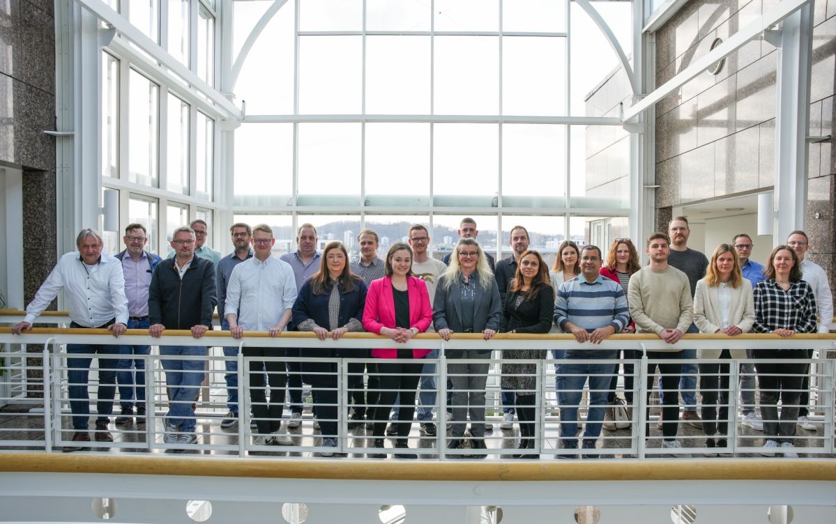24 Menschen stehen freundlich lächelnd auf einer Balustrade. Im Hintergrund ist ein großes Fenster mit einer verschwommenen urbanen Landschaft zu erkennen.