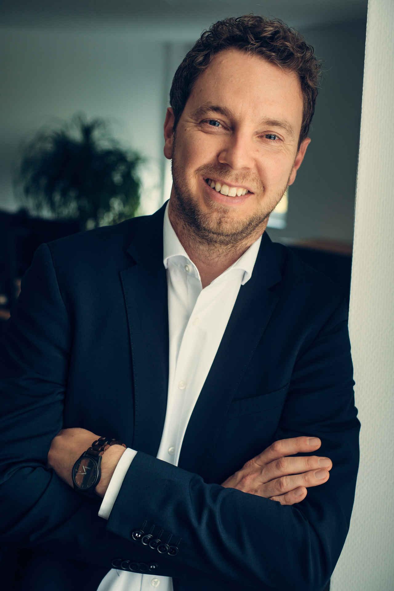 Ein professionell gekleideter Mann lehnt mit verschränkten Armen an einer Wand und lächelt freundlich.