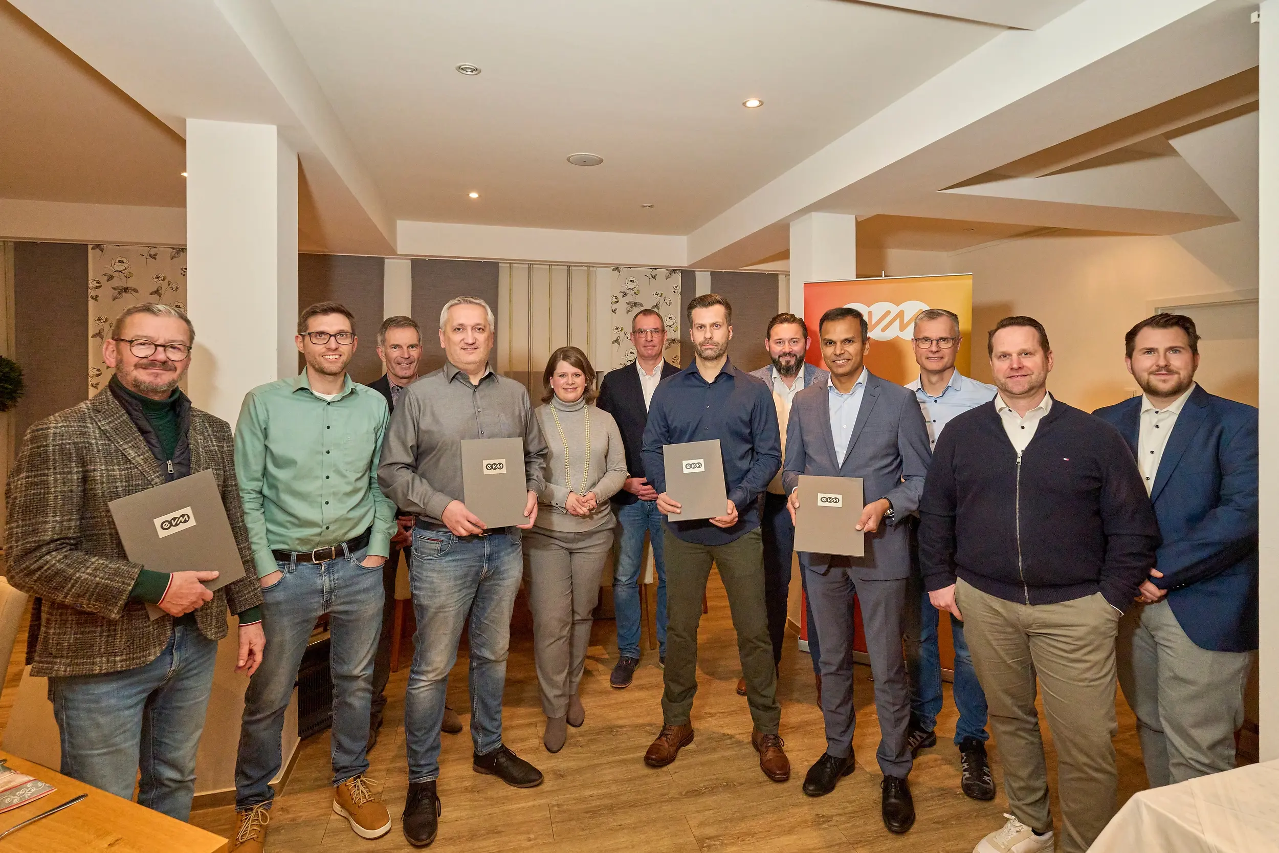 Eine Gruppe von zwölf Personen posiert lächelnd in einem Raum mit Holzboden, während vier Personen Urkunden halten. Die Atmosphäre wirkt freundlich.