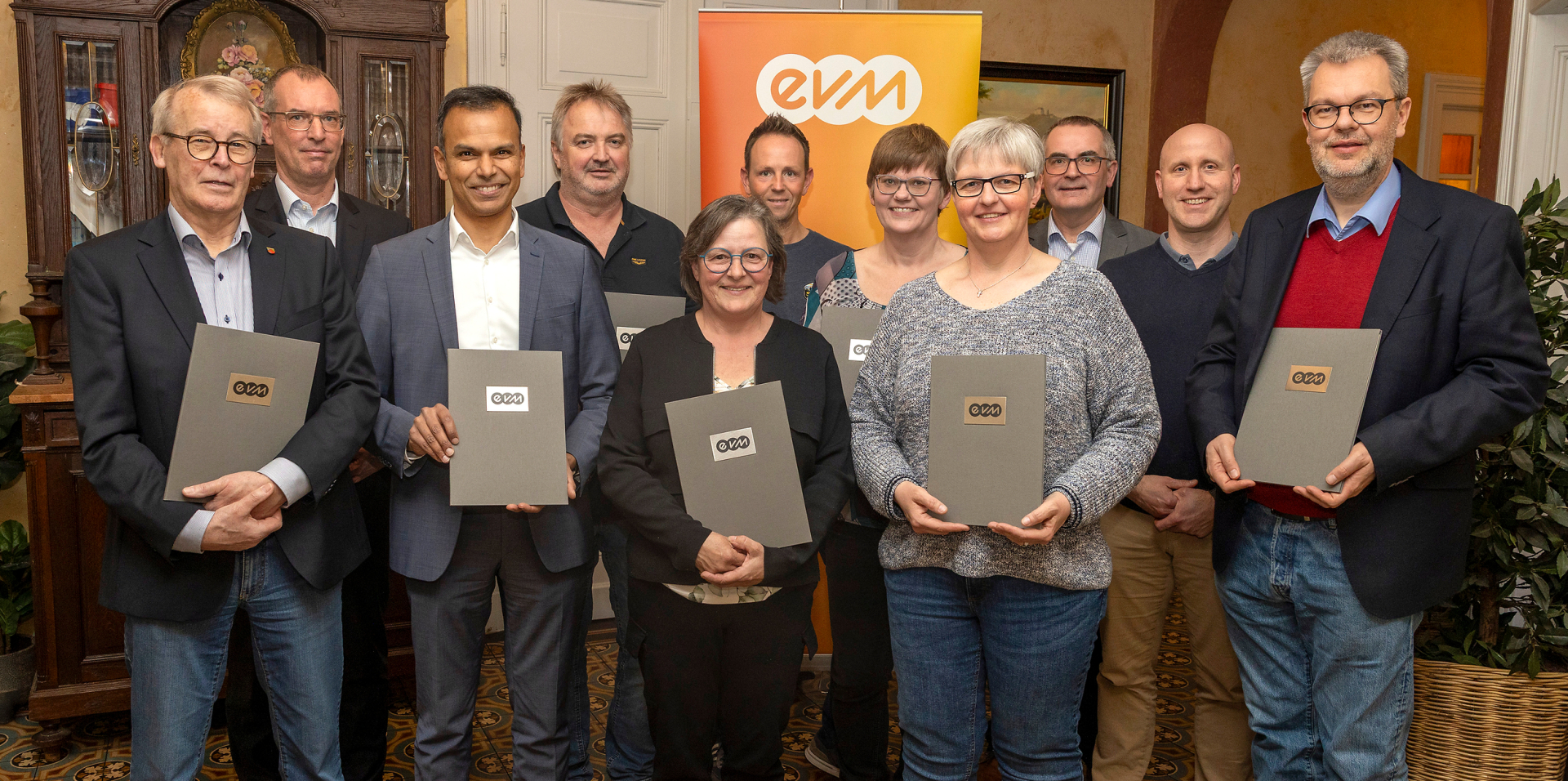 11 Menschen stehen vor einem orangenem Plakat mit der Aufschrift "evm" und lächeln freundlich in die Kamera. Sie sind professionell gekleidet und halten Mappen auf denen das evm-Logo zu erkennen ist.