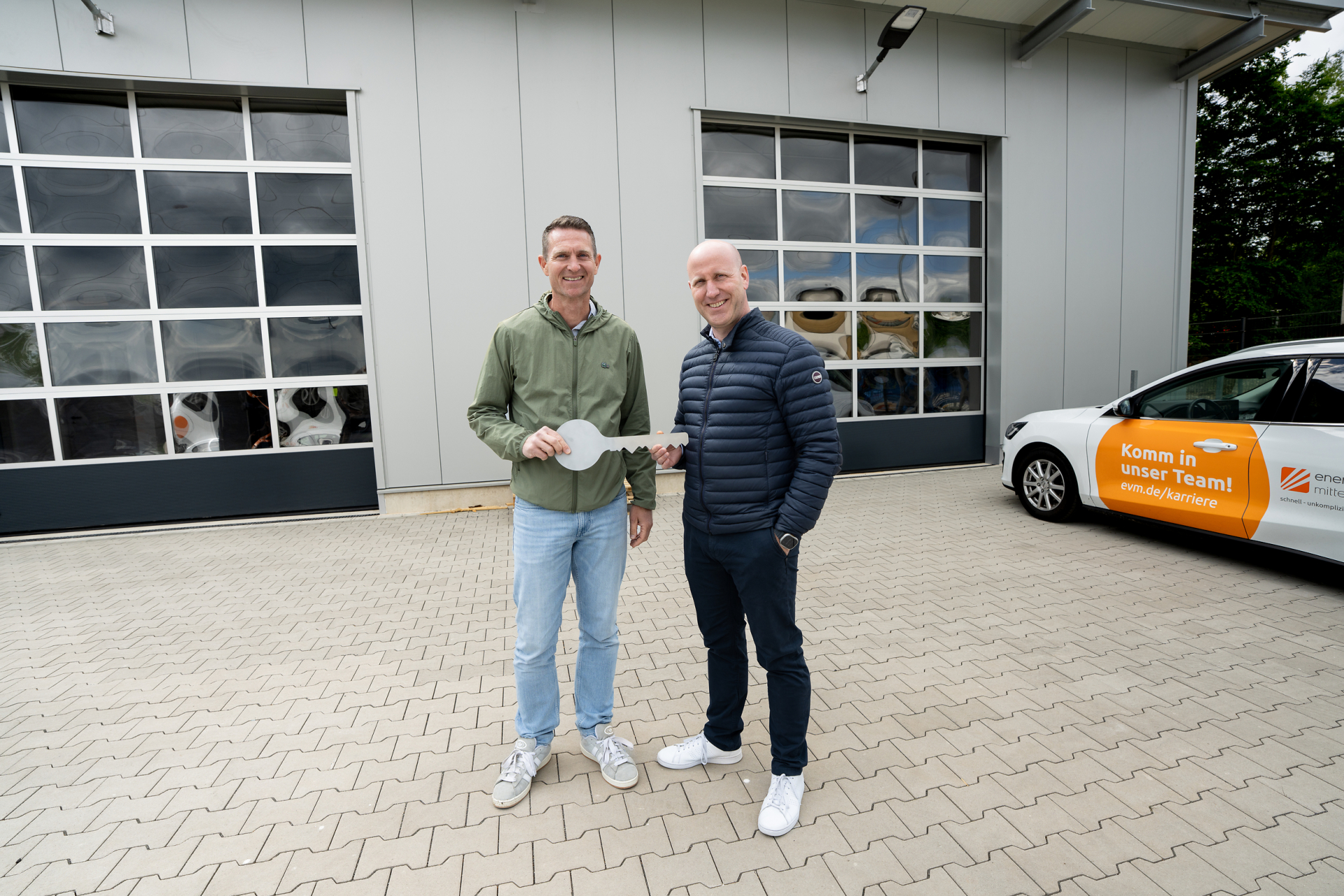 Zwei Männer stehen vor einer Lagerhalle und halten einen großen Schlüssel.