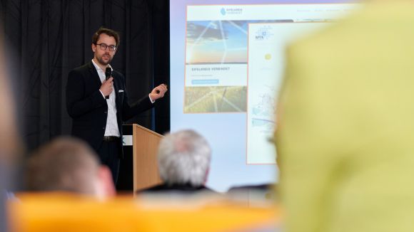 Ein Mann steht mit Mikrofon an einem Rednerpult, präsentiert Folien unter dem Titel "Beispiele Rheinland-Pfalz" in einem Konferenzraum. Zuschauer sind im Vordergrund sichtbar.
