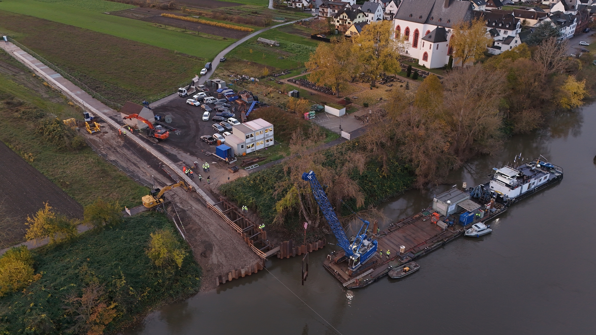 Himmelblick auf eine Baustelle die ins Wasser führt. Ein blauer Bagger steht auf einem Ponton.