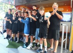 Eine Gruppe von Jungen in schwarzen Sporttrikots hält Pokale und einen Fußball, während sie posiert. Sie stehen lachend in einem sportlichen Veranstaltungsbereich in einem Zelt.