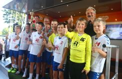 Eine Gruppe von Kindern in Sportkleidung steht lächelnd auf einer Bühne, hält eine Trophäe hoch und zeigt Daumen hoch. Erwachsene begleiten sie.