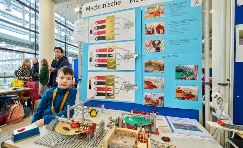 250210SDI1105 Ein junge steht neben einem Modell einer mechanischen Motorsteuerung und lächelt freundlich.