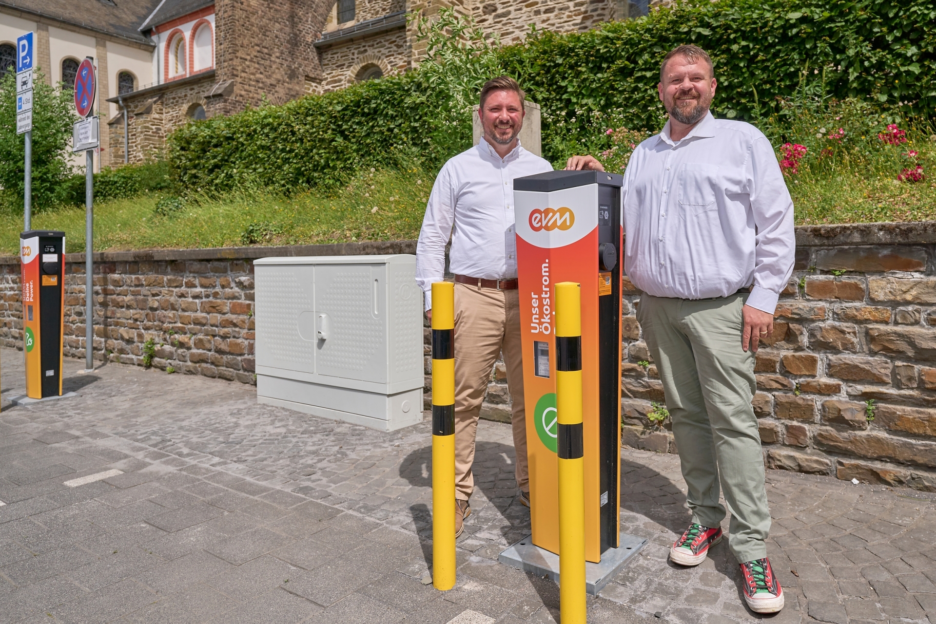 Zwei Männer stehen vor einer orangenen evm-Ladesäule und lächeln freundlich.