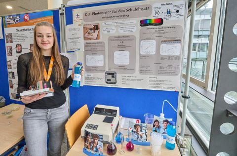250210SDI1080 Ein Mädchen steht vor einem Photometer und hält eine Falsche mit blauer Flüssigkeit in der Hand. Sie lächelt freundlich.