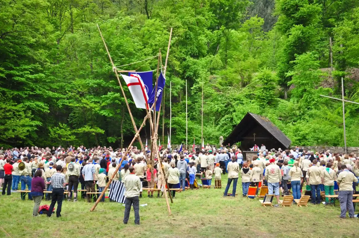 Eine Menschenmenge, größtenteils in beiger Pfadfinderkleidung gekleidet, versammelt sich auf einer Wiese. Fahnen sind an hohen Stangen befestigt, umgeben von üppigem Wald und einer hölzernen Bühne.