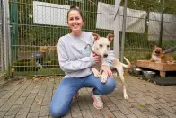 Eine Frau kniet neben einem hellbraunen Hund auf einem gepflasterten Boden und lächelt. Im Hintergrund ist ein eingezäunter Bereich mit einem weiteren Hund neben Futternäpfen sichtbar.