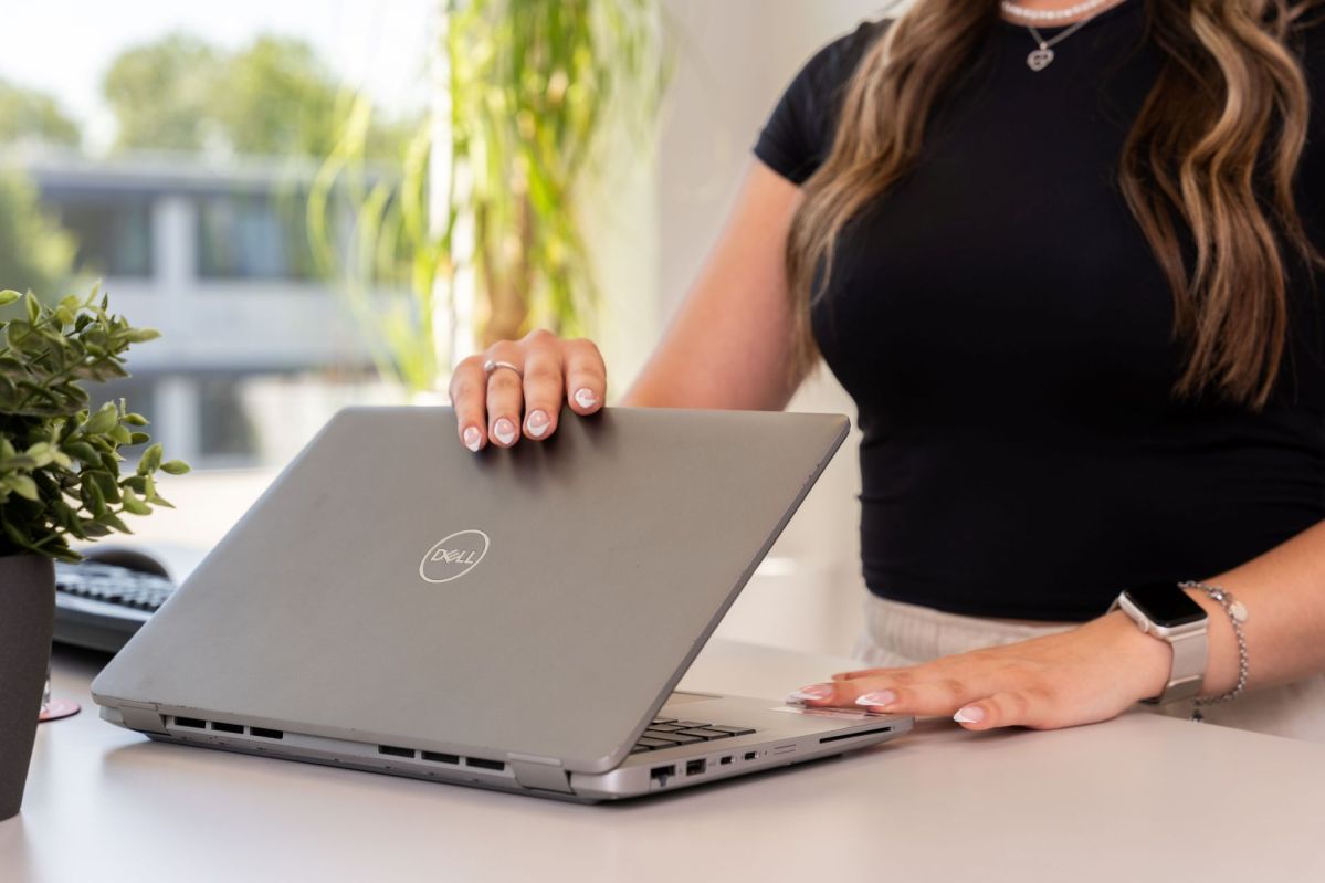 Eine junge nicht sichtbare Frau klappt einen grauen Computer auf. Der Computer steht auf einem modernen Schreibtisch in einem Büro.