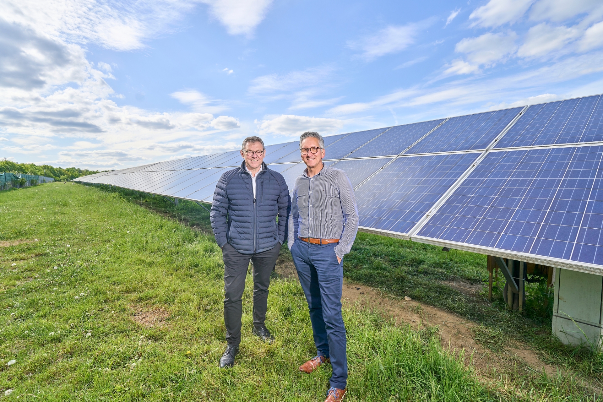 2 Männer stehen vor einem Solarpark und lächeln freundlich.
