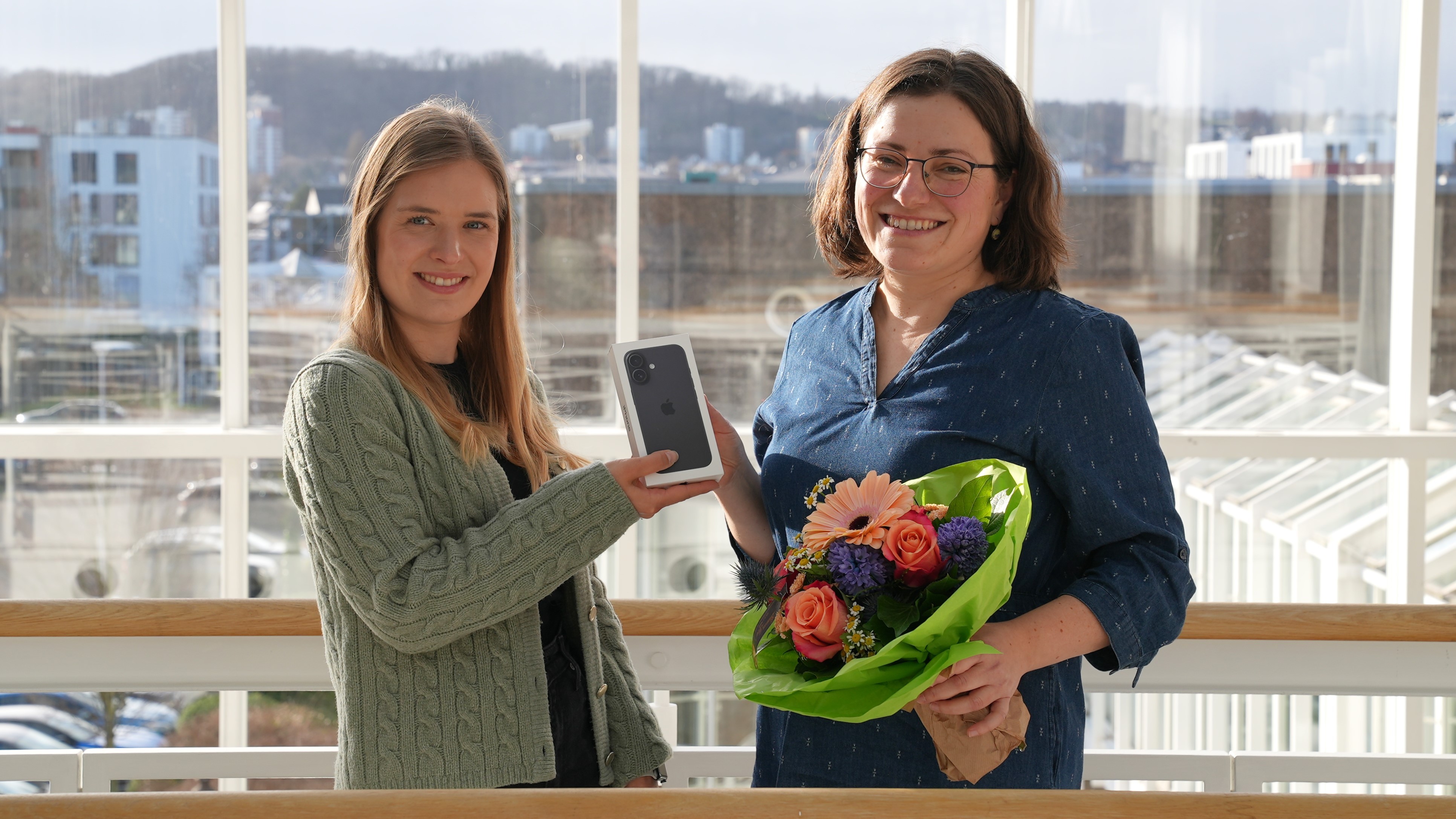 Zwei Frauen stehen auf einer Balustrade und lächeln freundlich. Die linke Frau hält ein Iphone was sie der rechten Frau übergibt die einen Blumenstrauß hält.