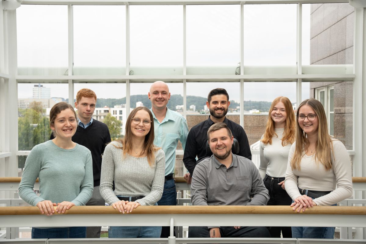 WdA-24 Vier junge Frauen und vier junge Männer stehen auf einer Balustrade vor einem verschwommenen urbanen Hintergrund und lächeln freundlich.