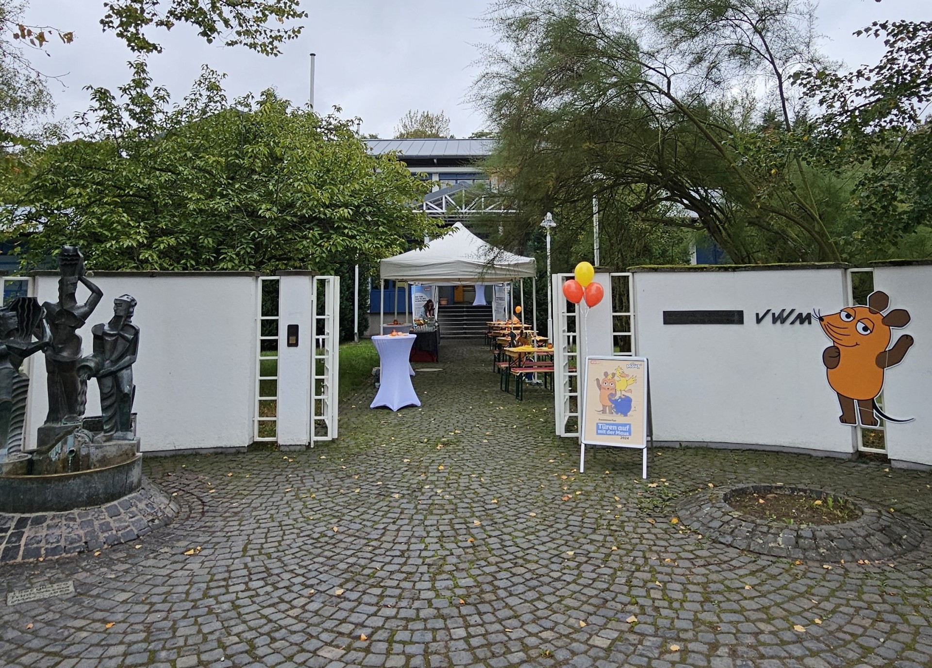 Der Eingang vom Wasserwerk Oberwerth. Eine weiße Wand mit einem Tor, daneben ein kleiner Brunnen. Rechts ist die orangene Maus von Sendung mit der Maus abgebildet. Im Eingang befinden sich Tische und orangene Ballons.