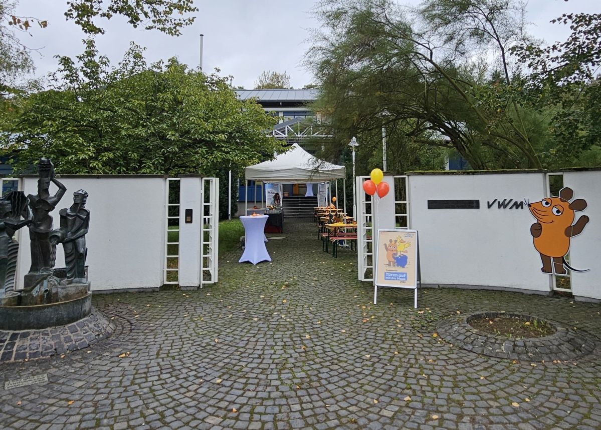 20241003_093449 Der Eingang vom Wasserwerk Oberwerth. Eine weiße Wand mit einem Tor, daneben ein kleiner Brunnen. Rechts ist die orangene Maus von Sendung mit der Maus abgebildet. Im Eingang befinden sich Tische und orangene Ballons.