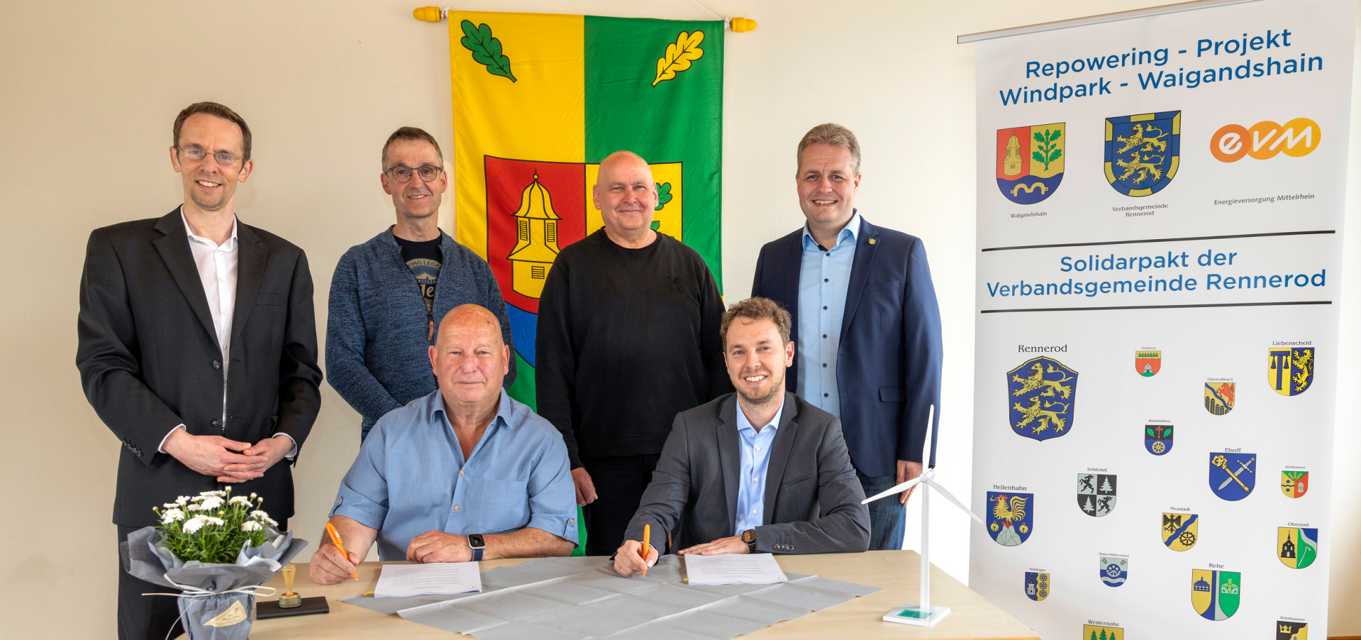 Zwei Männer sitzen an einem Tisch und unterschreiben ein Dokument. Hinter ihnen stehen vier Männer und lächeln freundlich.