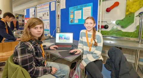 250210SDI1098 Zwei Mädchen sitzen an einem Tisch auf dem ein Computer steht und lächeln freundlich.