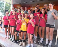 Eine Gruppe junger Menschen in roten Sporttrikots hält einen goldenen Pokal, lächelt und zeigt Daumen hoch. Sie stehen zusammen auf einer Bühne im Freien bei sonnigem Wetter.