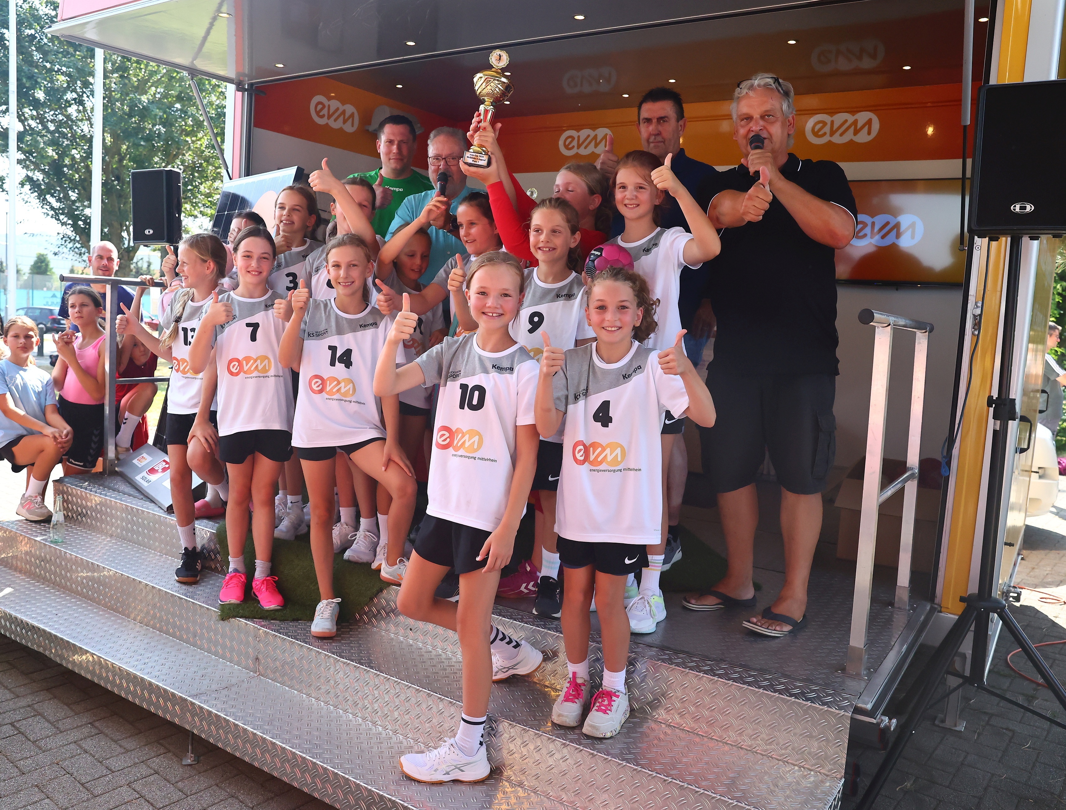 Eine Gruppe von Mädchen in Sportbekleidung posiert mit einem Pokal auf einer Bühne. Im Hintergrund stehen Erwachsene und es gibt ein EVM-Banner. Zuschauer sitzen und stehen am Rand.