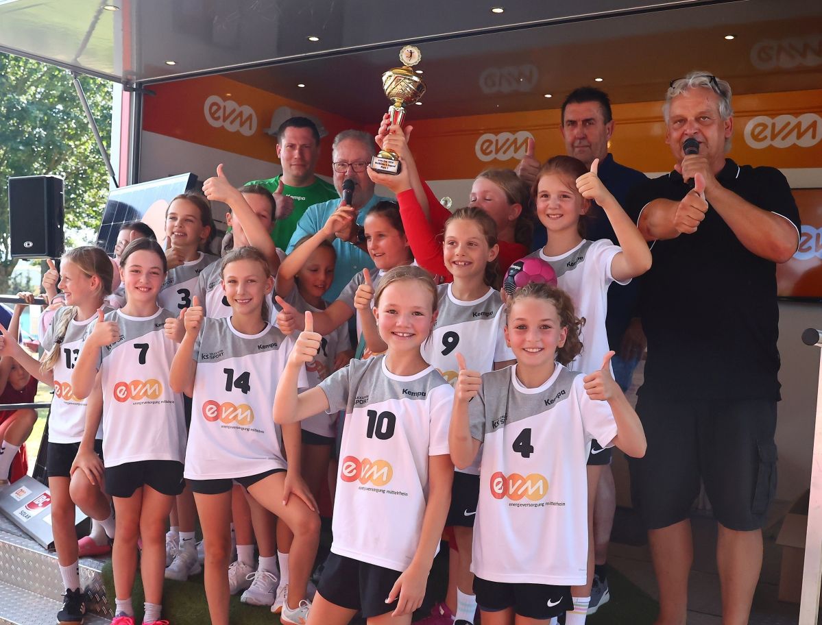 Eine Gruppe von Mädchen in Sportbekleidung posiert mit einem Pokal auf einer Bühne. Im Hintergrund stehen Erwachsene und es gibt ein EVM-Banner. Zuschauer sitzen und stehen am Rand.