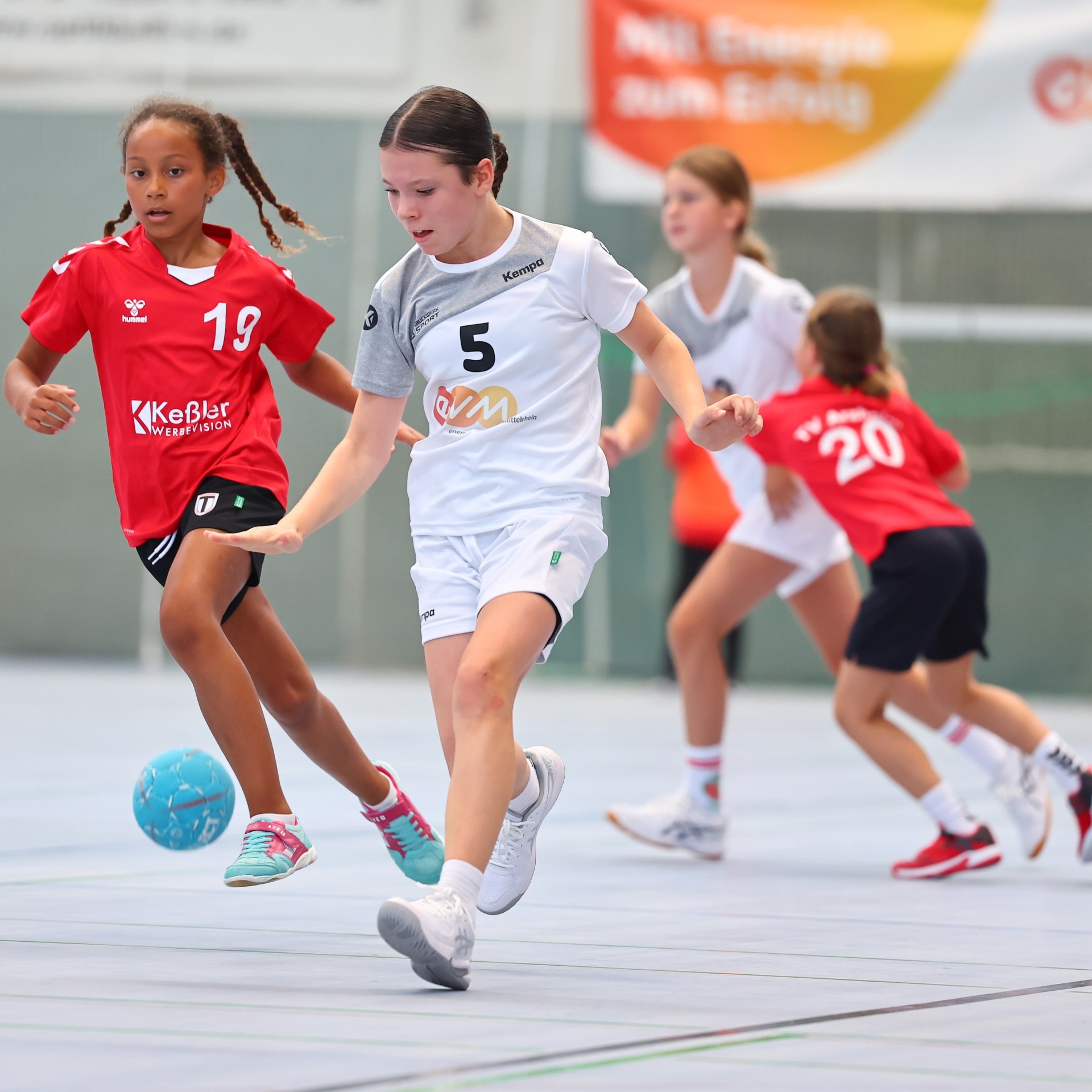 Vier Mädchen in roten Trikots und weißen evm Trikots spielen Handball in einer Sporthalle.